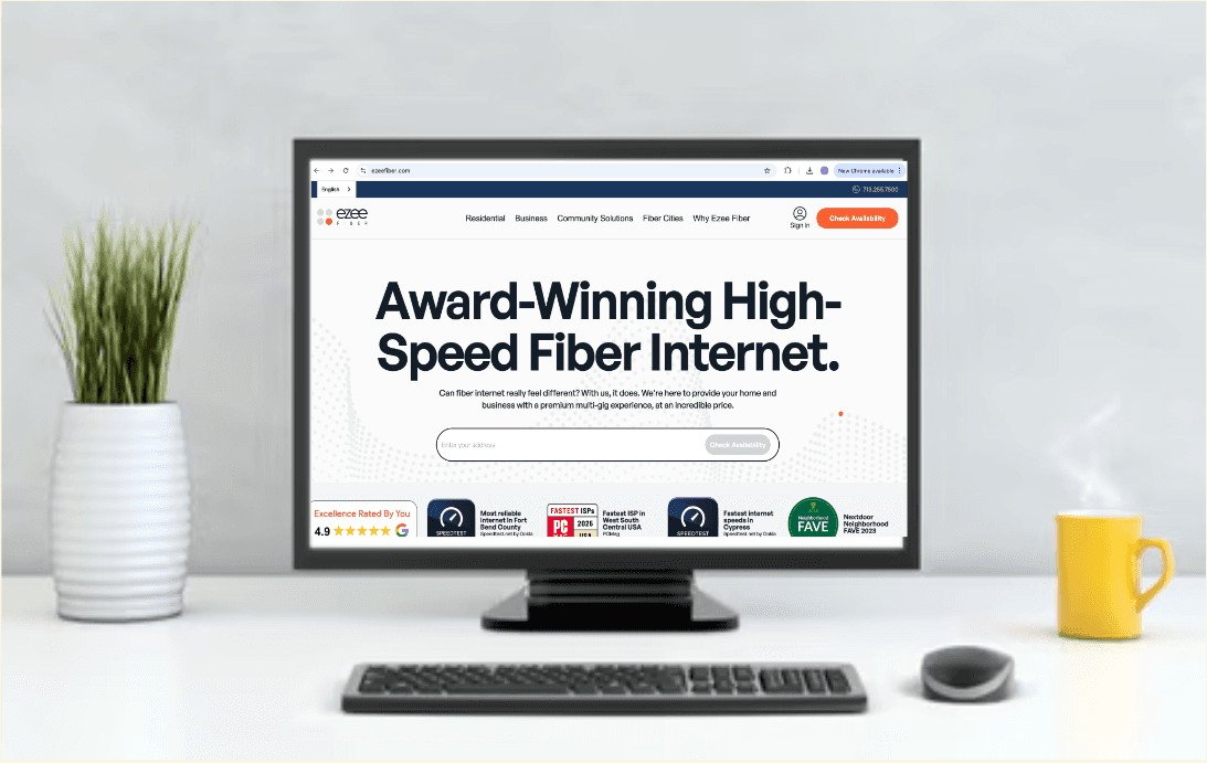 A computer monitor on a white desk displays a website promoting award-winning high-speed fiber internet for EzeeFiber. A potted plant and a yellow mug are placed on either side of the monitor.